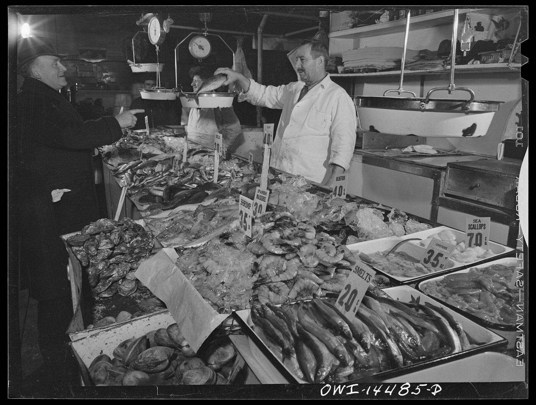 Fotografia d’archivio in bianco e nero: un uomo con il grembiule dietro un banco di pesce ghiacciato e cartellini scritti a mano