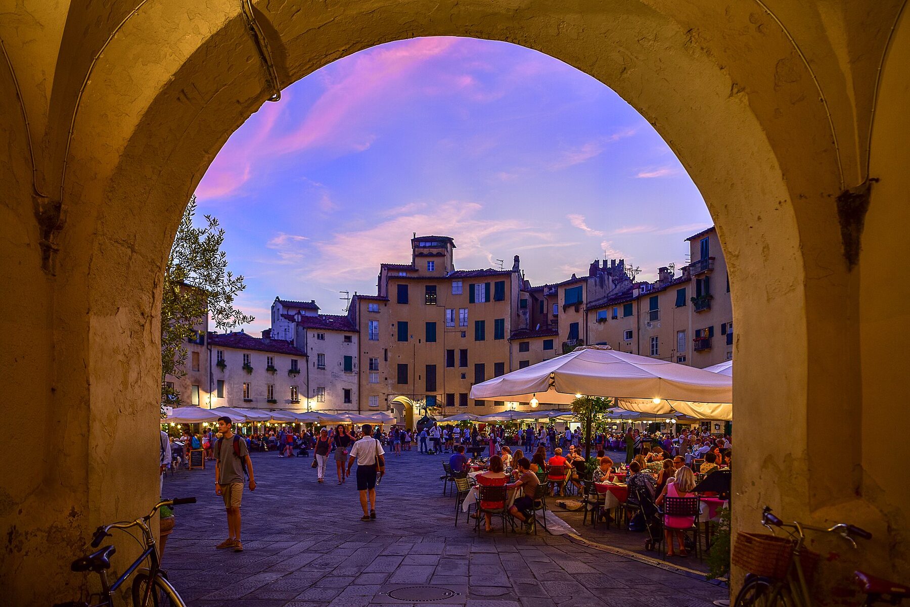 Tavolini e case color pastello visti attraverso l’arco di pietra di Piazza dell’Anfiteatro a Lucca, al tramonto